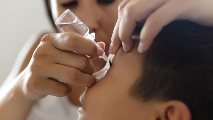 A photo of a boy with an eye drop bottle being held above his eye by his mom in the background.