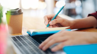 a close-up photo of a desk. One hand on a computer keyboard while the other hand takes notes