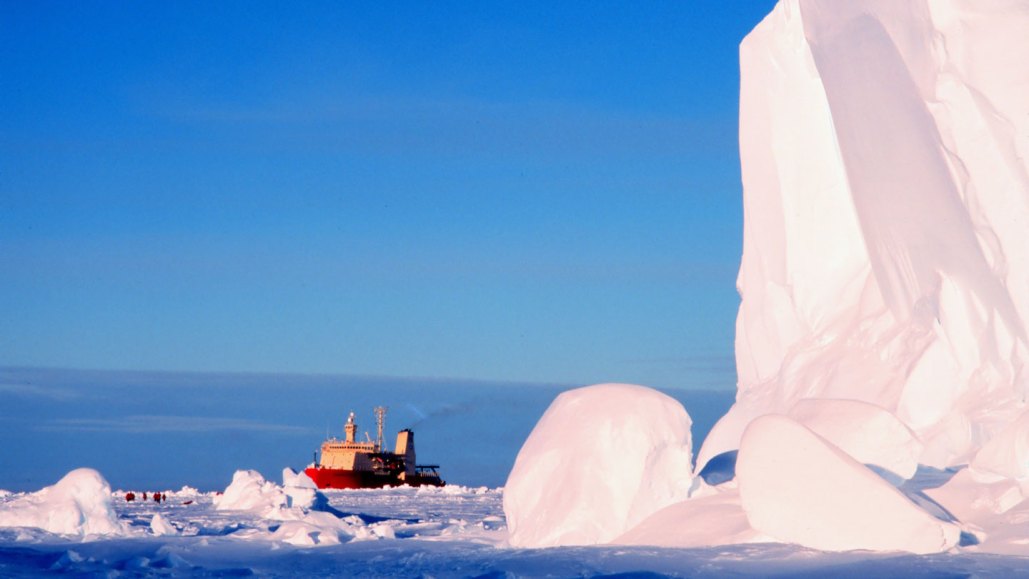 A massive ice cliff towers over a boat sailing in ice-encrusted waters in the background