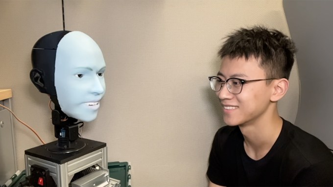 On the left, Emo, a robot with a blue silicone face, smiles in tandem with researcher Yuhang Hu, on the right. Hu wears a black t-shirt.