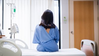 A woman with black hair sits on a white hospital bed with her back to the camera. She is wearing a blue hospital gown. Now, people at hospitals must obtain consent prior to intimate exams.