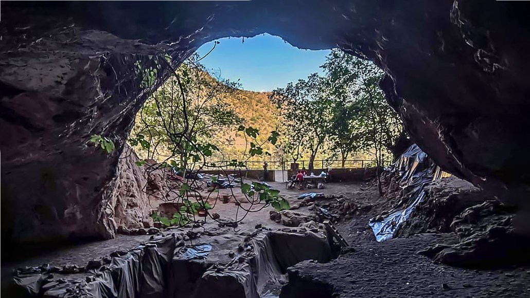 The view to the landscape from inside a cave in Morocco that archaeologists have been excavating. You can see trenches in the foreground and a few trees and a hillside with some vegetation beyond.