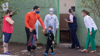 A man and woman wearing face masks greet a young boy walking out of a school where four people, also in face masks, are standing in the doorway