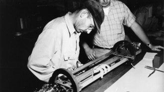 black and white photo of a man in a cap with glasses leaning over a tabletop device while another man looks on