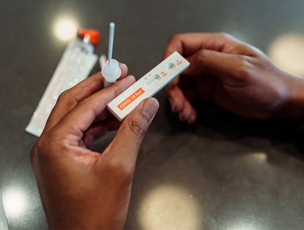 The photo shows a person's hands holding the bits and pieces of a home COVID test, including the swab and the test strip. No results have shown up yet.