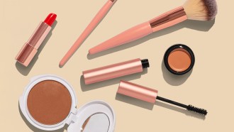 several makeup items - lipstick, a compact, blush brush, mascara - sit on a cream-colored background