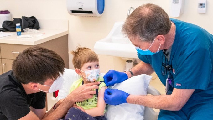 a child sits on a hospital bed as a parent holds his hand and a doctor puts a band aid on his shoulder