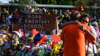 Two people hug outside a sign that reads ROBB ELEMENTARY SCHOOL. There are flowers all around the sign.