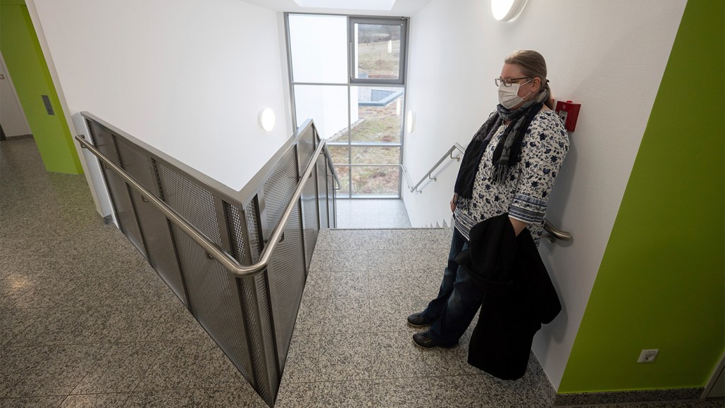 A woman with long COVID rests against a white wall in a stairwell. She is wearing glasses and a white mask.