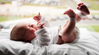 A photo of a crying baby laying on her back on a pillow in front of a window.