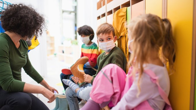 A teacher wearing a mask looks to several young children, also wearing masks and leaning against a cupboard