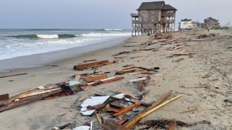 Debris from a collapsed house litters a beach in North Carolina. In the distance, a house on stilts still stands at the very edge of the ocean.