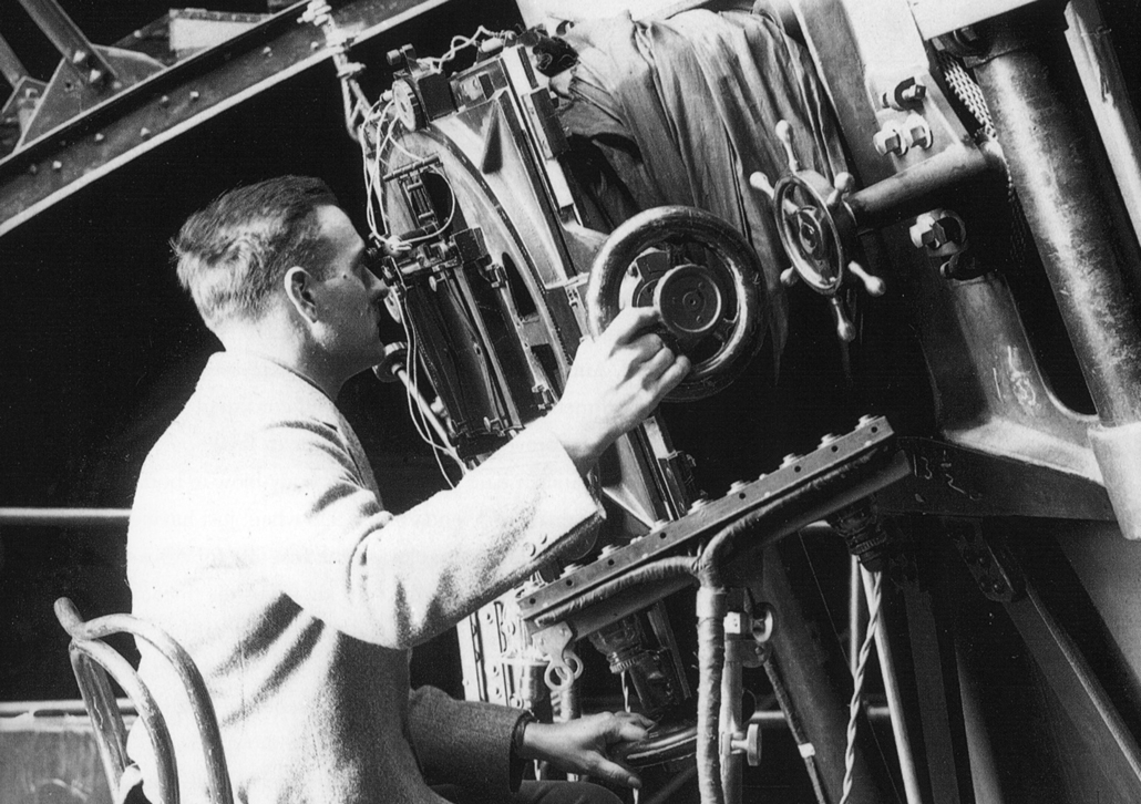 Black and white photo of Edwin Hubble looking into an old telescope