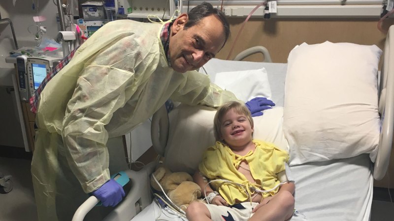 Will Ungerer lies in a hospital bed, next to a teddy bear, as doctor Jerry Mendell leans over him. Both are looking at the camera and smiling.