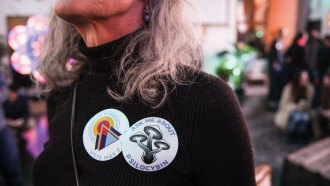 image of a woman wearing a sticker that reads "Ask me about psilocybin" and another sticker that reads "Decriminalize Mushrooms"