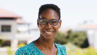 Hawa Racine Thiam smiles at the camera. She has short, dark hair and is wearing blue rimmed eyeglasses, a turquoise shirt and a white necklace.