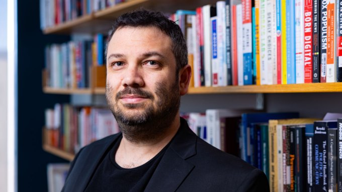 Yotam Ophir looks at the camera with a slight smile. He is standing in front of a bookcase full of books of different sizes and colors. Ophir has short, dark hair and has short beard and mustache.